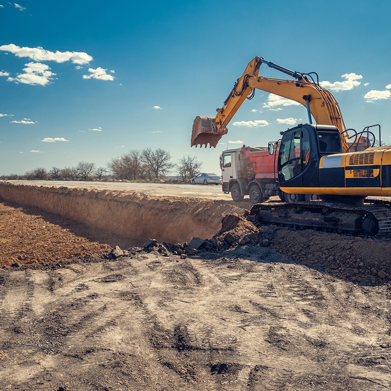 Excavatrice jaune creusant une tranchée profonde, chargeant une benne d'un camion-benne sous un ciel bleu clair.