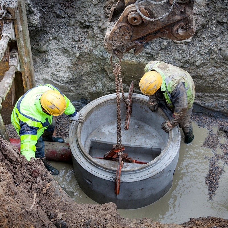 Deux ouvriers en tenue de sécurité jaune-vert et casques jaunes installent un anneau de chambre de visite en béton préfabriqué dans une tranchée boueuse, aidés par une grue.