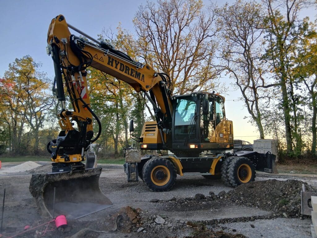 HYDREMA : L'excavatrice sur pneus idéale pour tous vos chantiers. Excavatrice sur pneus HYDREMA jaune travaillant sur un chantier. La machine creuse la terre avec son godet. Ciel bleu et arbres en arrière-plan.