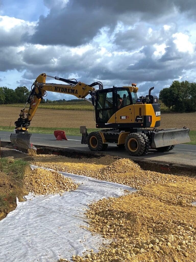 Pelle sur pneus HYDREMA MX14 en action sur chantier. Pelle HYDREMA MX14 creusant une tranchée. Matériaux (gravier sur géotextile) pour la fondation de travaux routiers sous ciel nuageux.