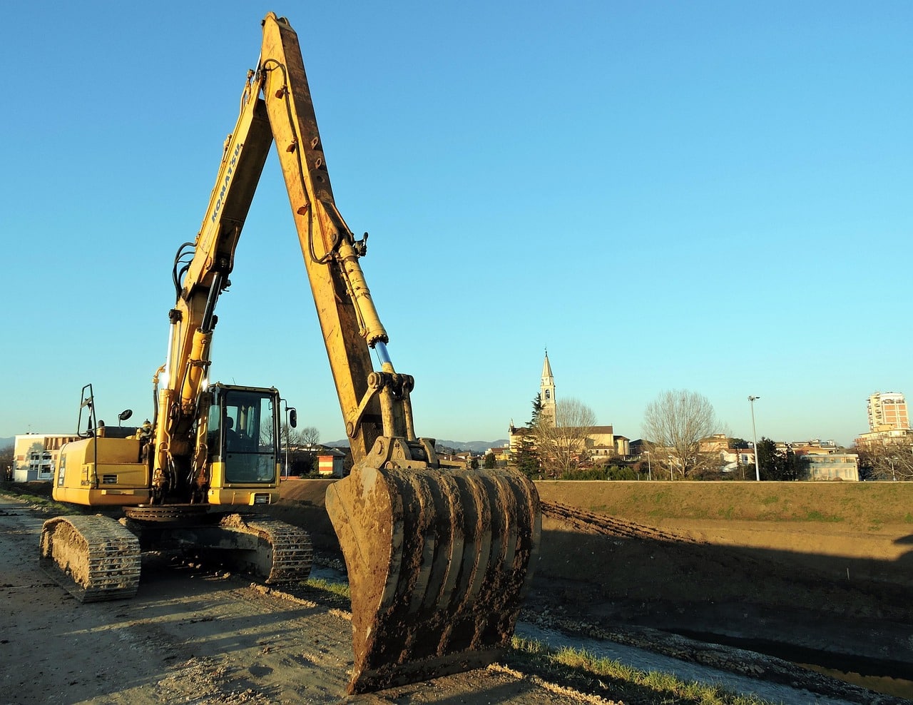 Excavation au Coucher du Soleil : Travaux Lourds et Lumière Dorée Godet d'excavatrice jaune déversant de la terre et des roches au coucher du soleil dans une carrière, créant une forte lueur dorée.