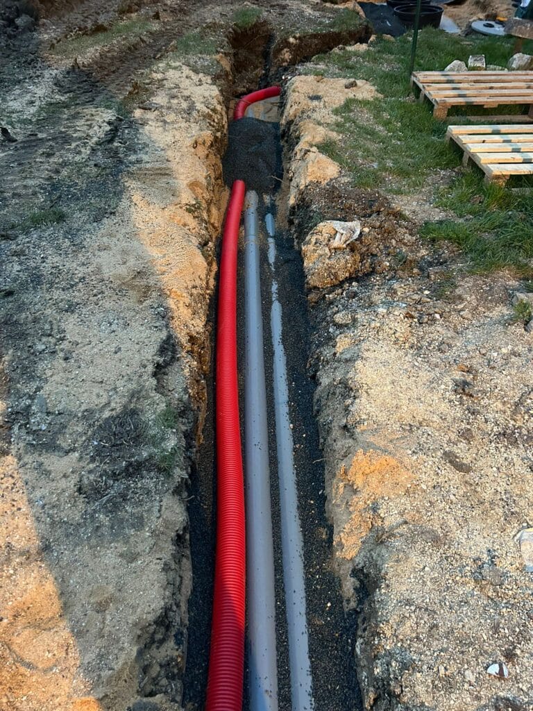 Pose de gaines et tuyaux en tranchée de terrassement Gaine rouge et tuyaux gris posés dans une tranchée sur lit de gravier, chantier extérieur.