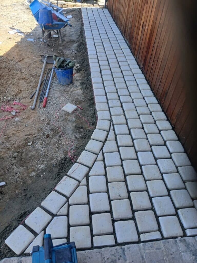 Pose de pavés extérieurs : aménagement d'allée de jardin Pose de pavés beiges pour une allée de jardin bordant une clôture en bois. Chantier extérieur en cours.